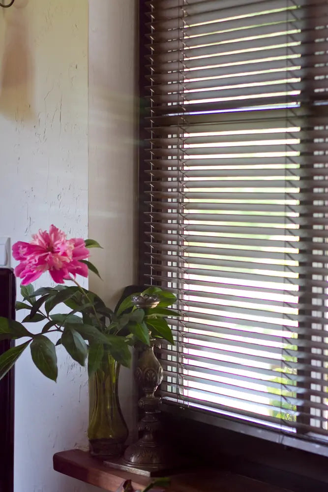 Mörkbrun träpersienn med 25 mm lameller vid fönster, med rosa blomma i vas på fönsterbrädan i mjukt dagsljus